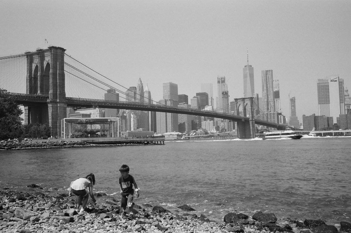 A black and white photo of two children playing in the water with a bridge in the background.