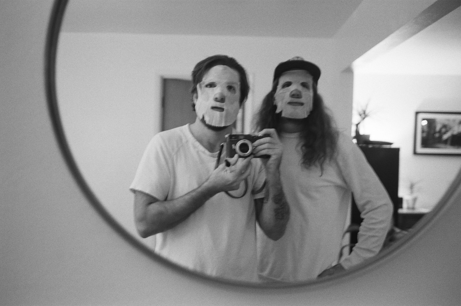 Two men wearing face masks are taking a picture of themselves in a mirror.