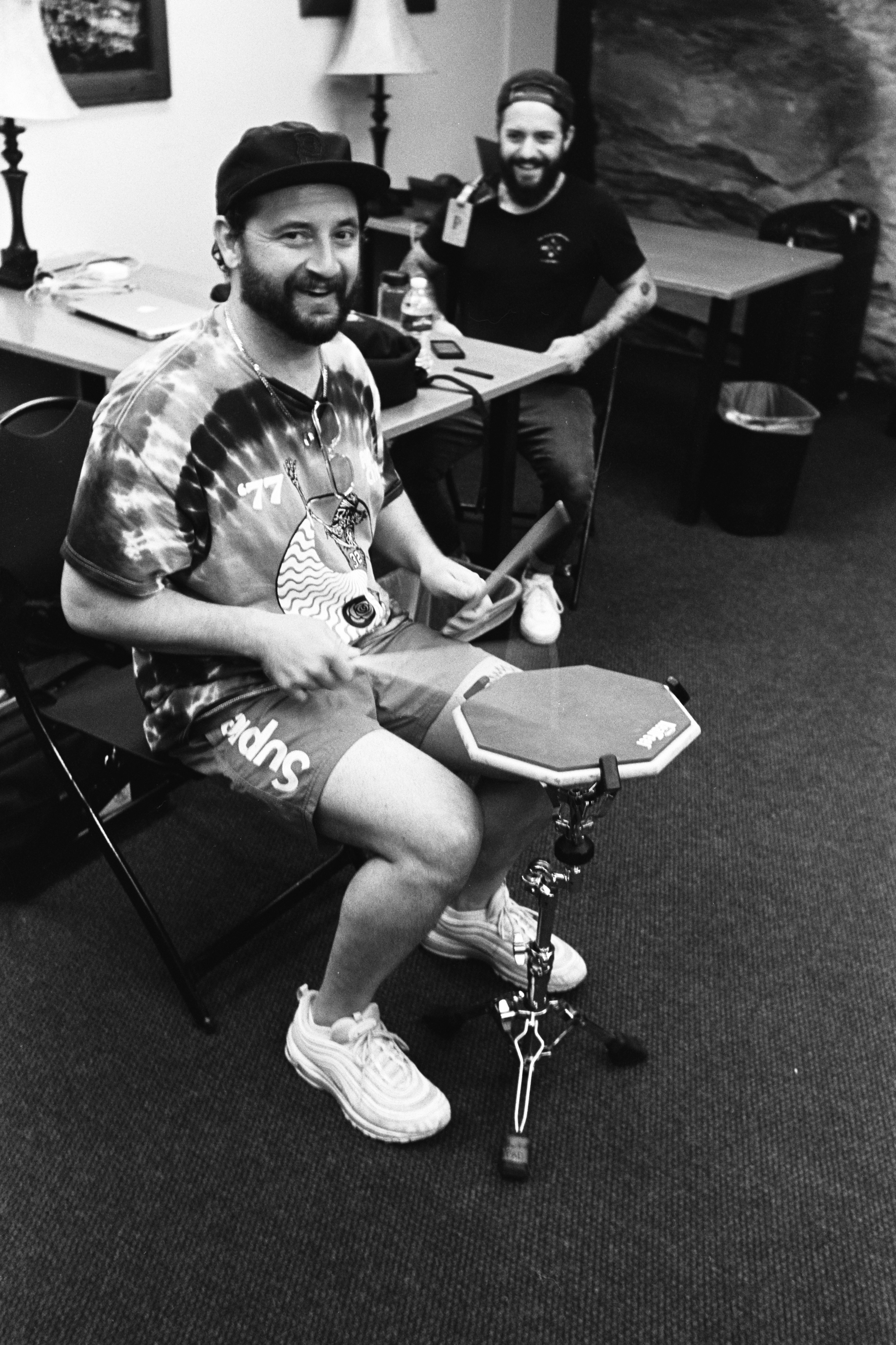 A man in a tie dye shirt is playing drums in a black and white photo.