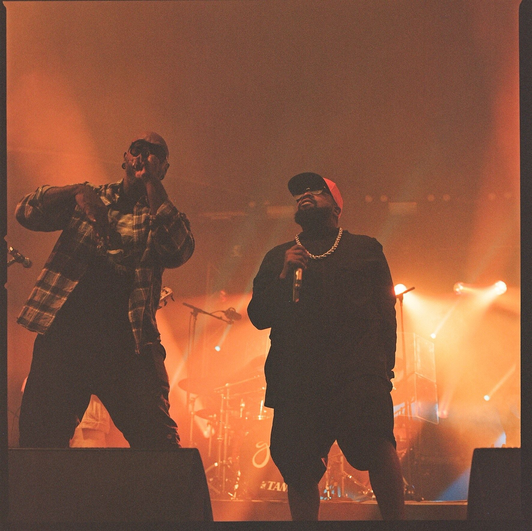 Two men singing into microphones on a stage with a drum set in the background