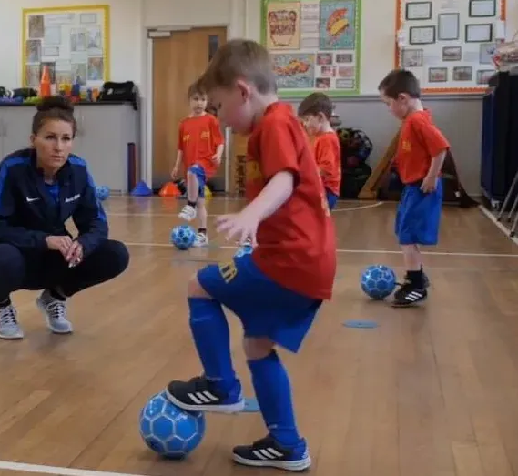A young boy in a red shirt balances a soccer ball with his foot during a sports lesson.