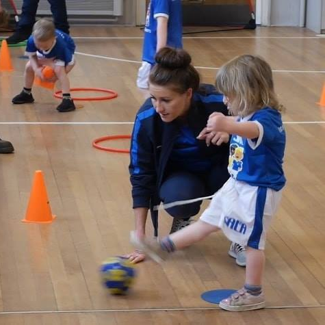 Woman helps a young child kick a soccer ball during a sports class in a gym.
