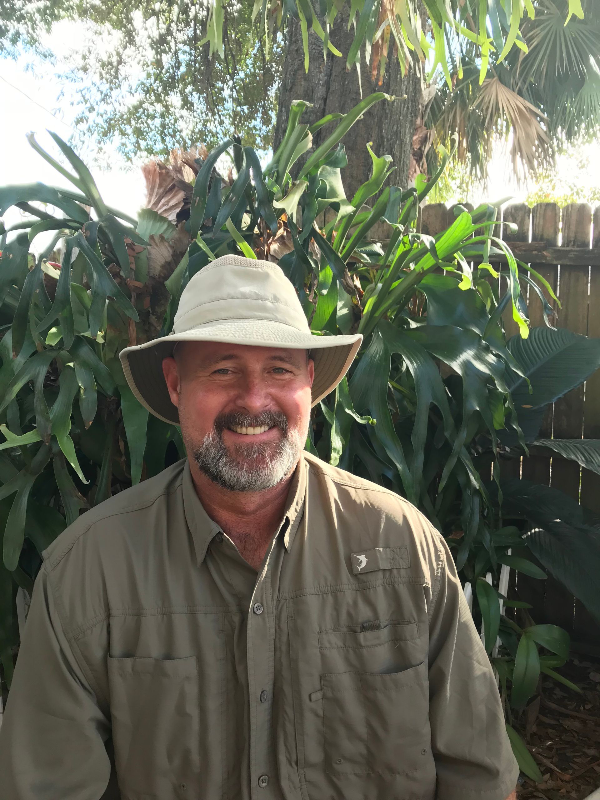 Man in orange shirt, hat, and sunglasses with a blue and white neck gaiter, smiling in front of buildings and blue sky.