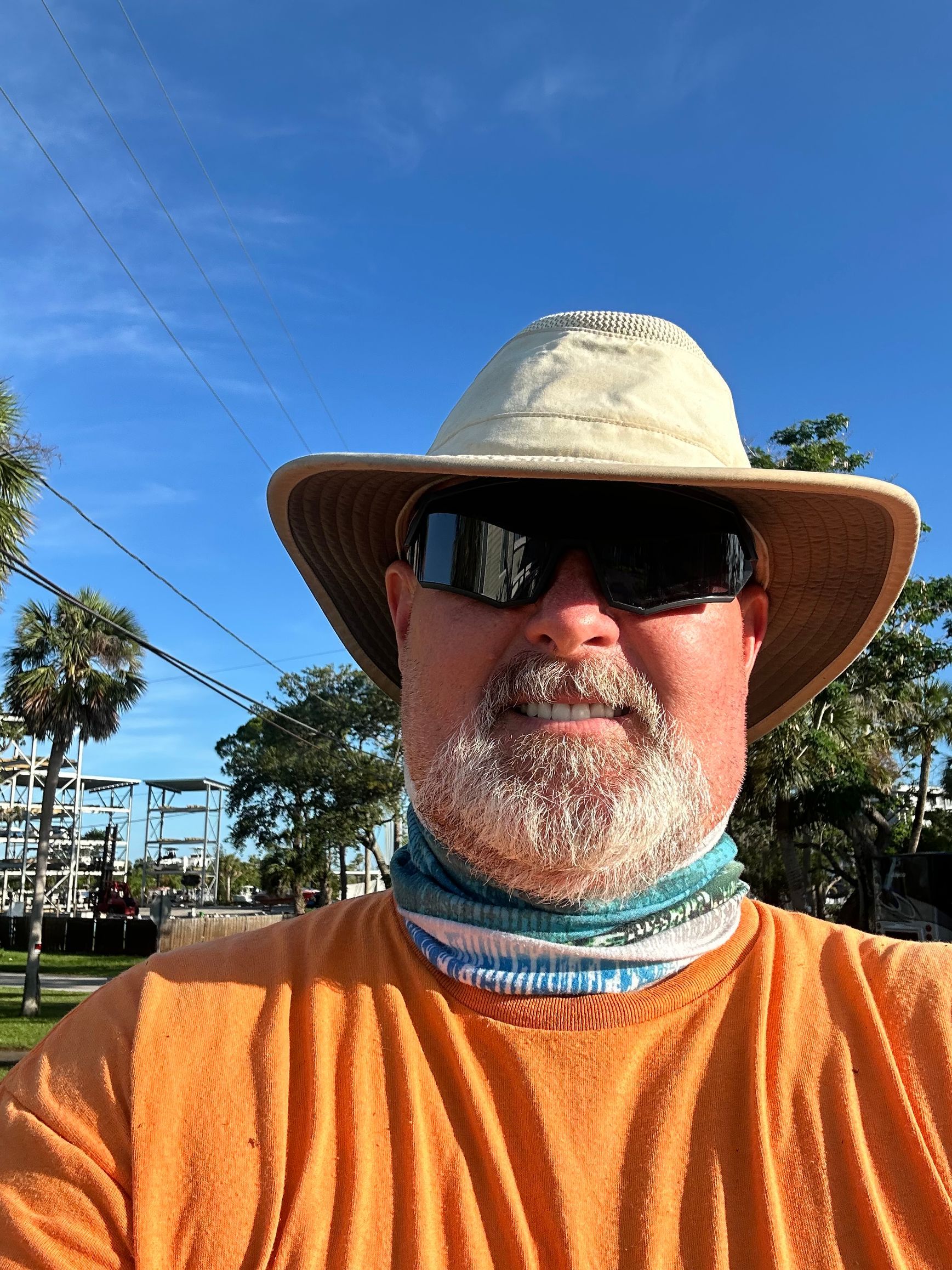 Man in orange shirt, hat, and sunglasses with a blue and white neck gaiter, smiling in front of buildings and blue sky.