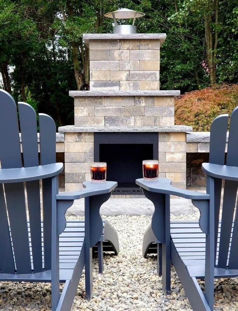Two gray Adirondack chairs face a stone fireplace with drinks, outdoor setting.