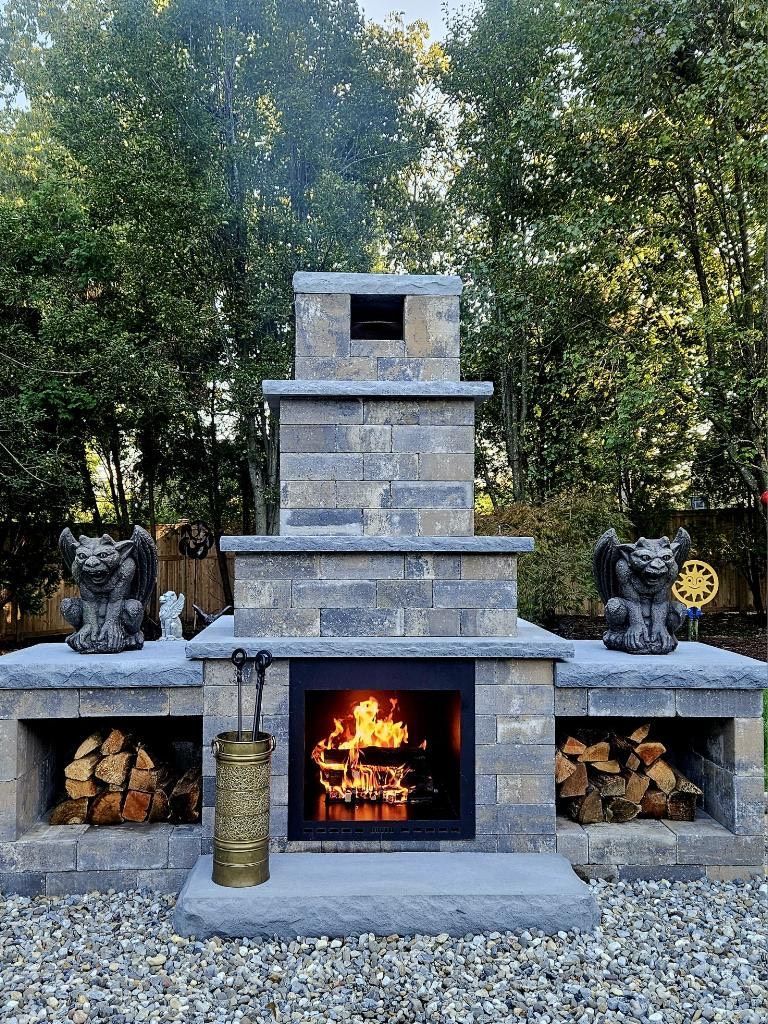 Outdoor fireplace with burning fire and stacked firewood. Two gargoyles sit on either side.