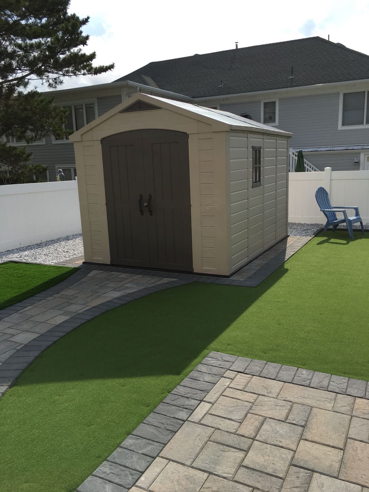 Tan and brown storage shed with a pathway and lawn. A blue chair sits nearby.