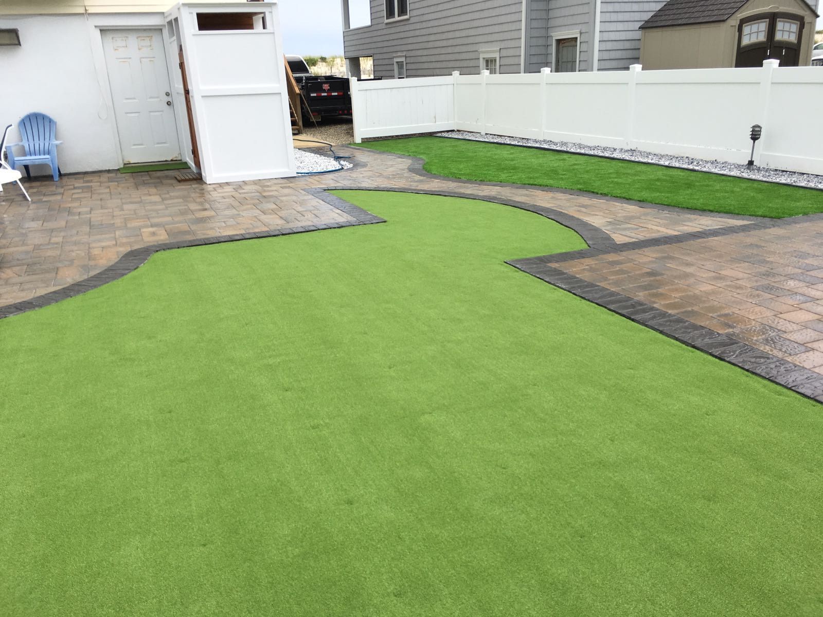 Backyard with artificial green turf bordered by patterned concrete and a white fence.
