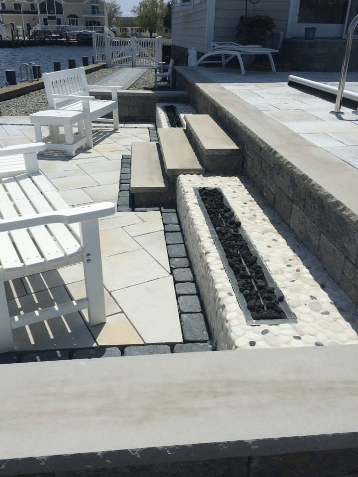 Outdoor patio with white furniture, built-in benches, and a fire pit, next to water.