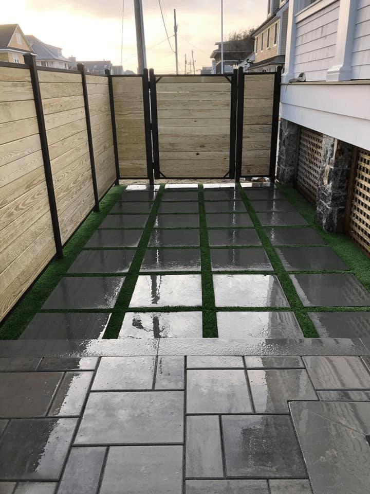 Stone patio with wet pavers, green turf, and wooden fence.
