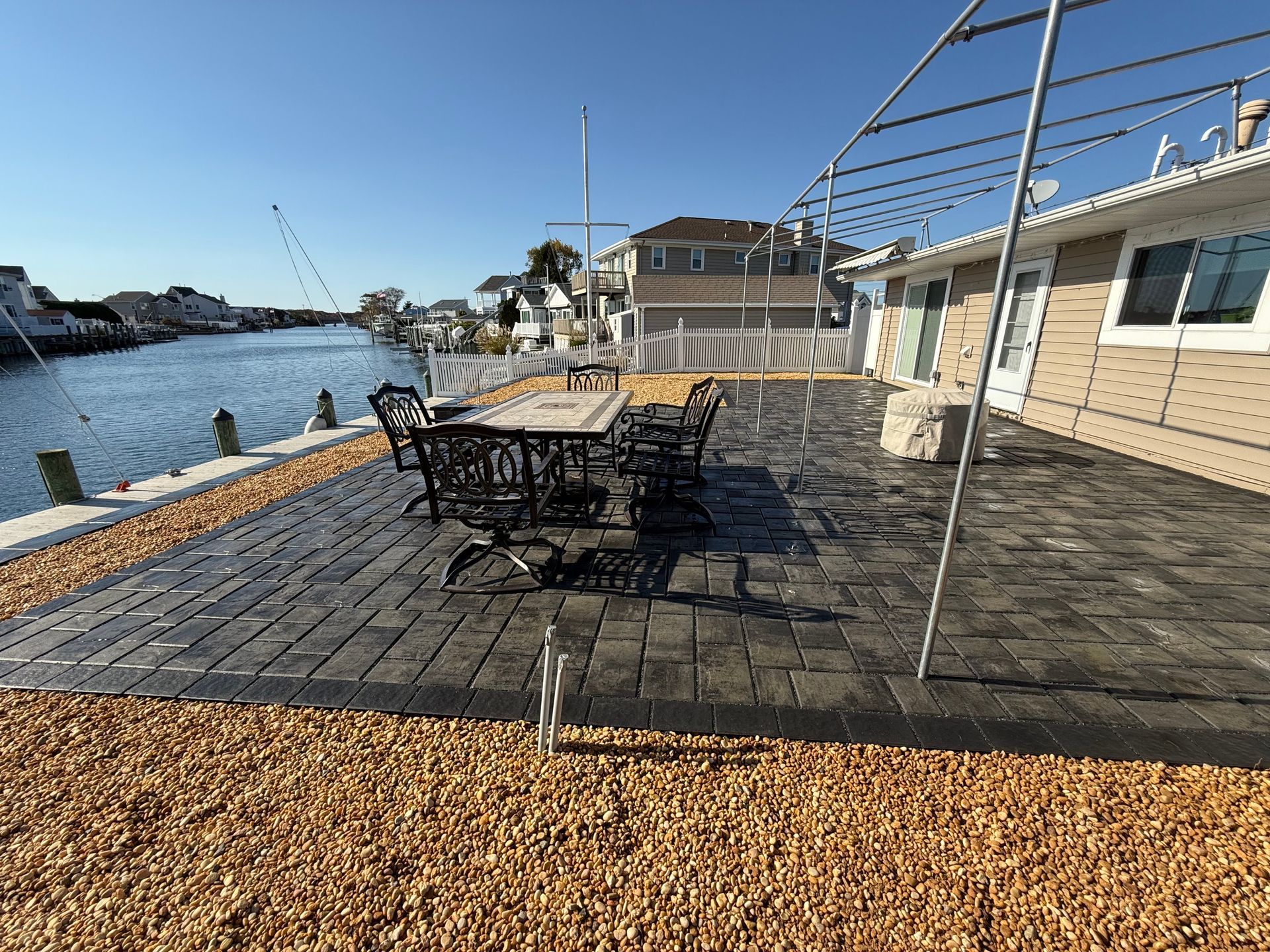 Patio overlooking water, with seating, table, and house in the background. Sunny day.