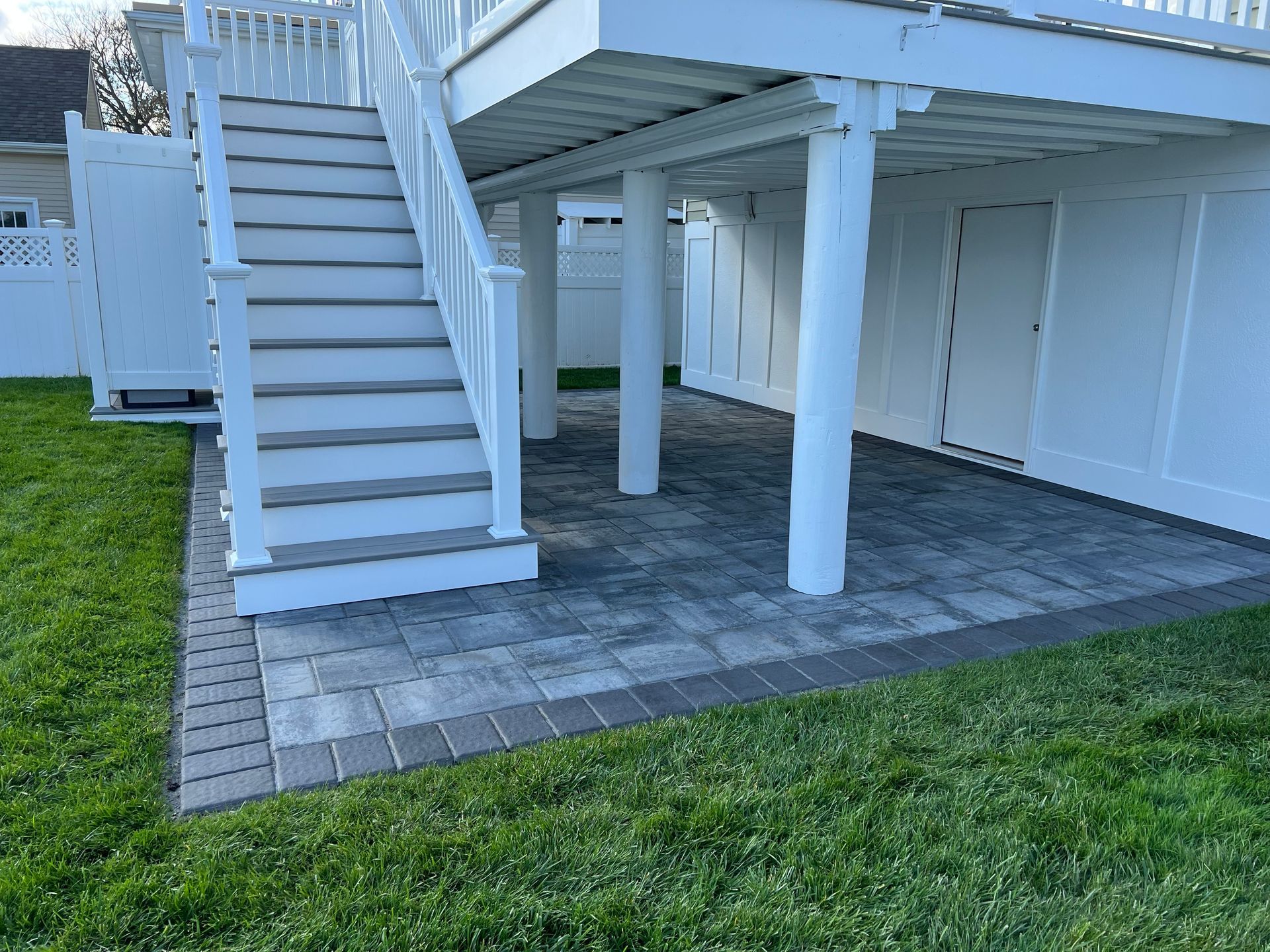 Stone patio under a white deck with stairs, surrounded by green grass and a white fence.