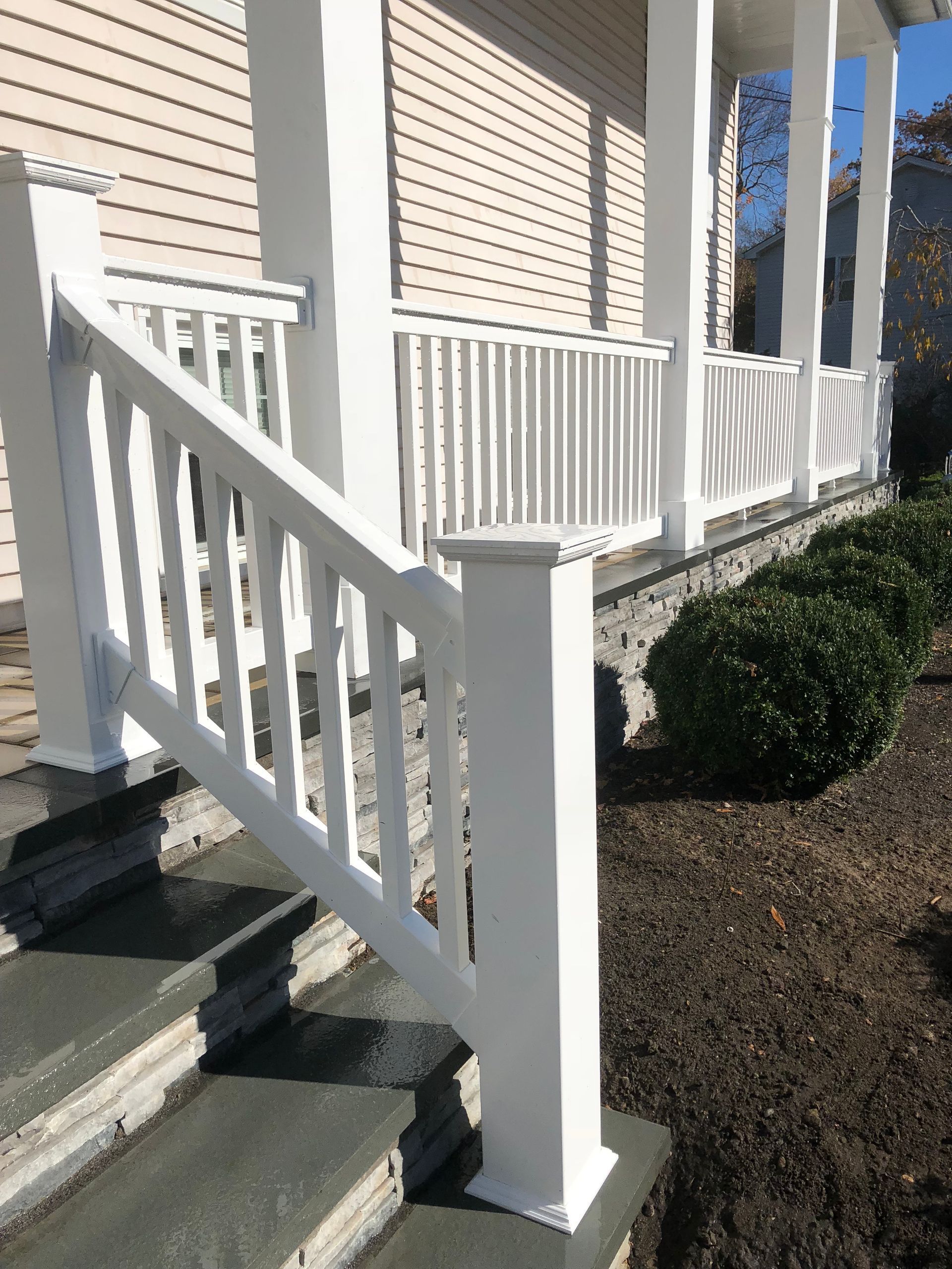 White porch with steps, railing, and columns. Beige siding, green shrubs, and brown mulch.