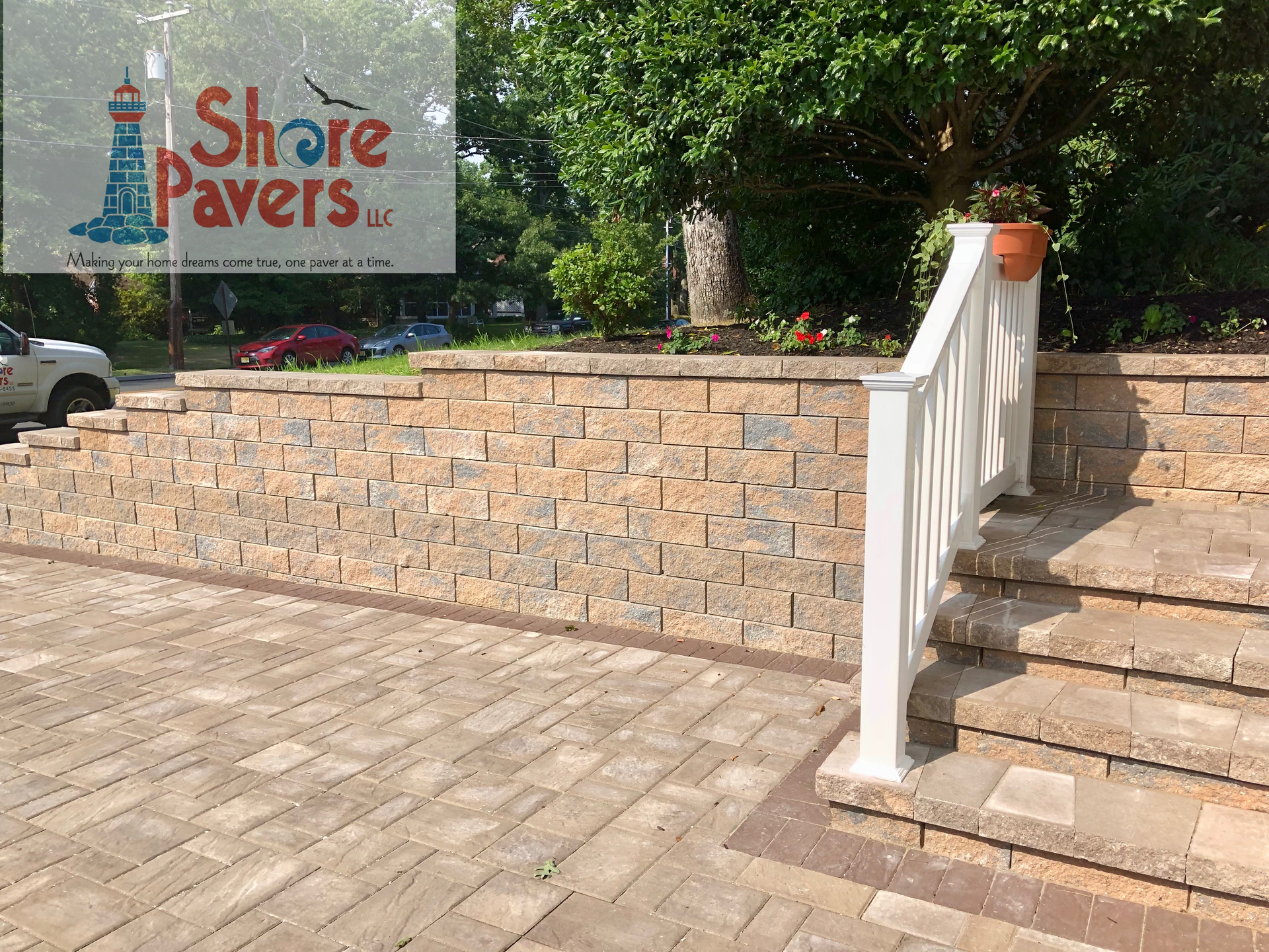 Brick retaining wall with stairs and handrail. Driveway paved with bricks, trees in background.