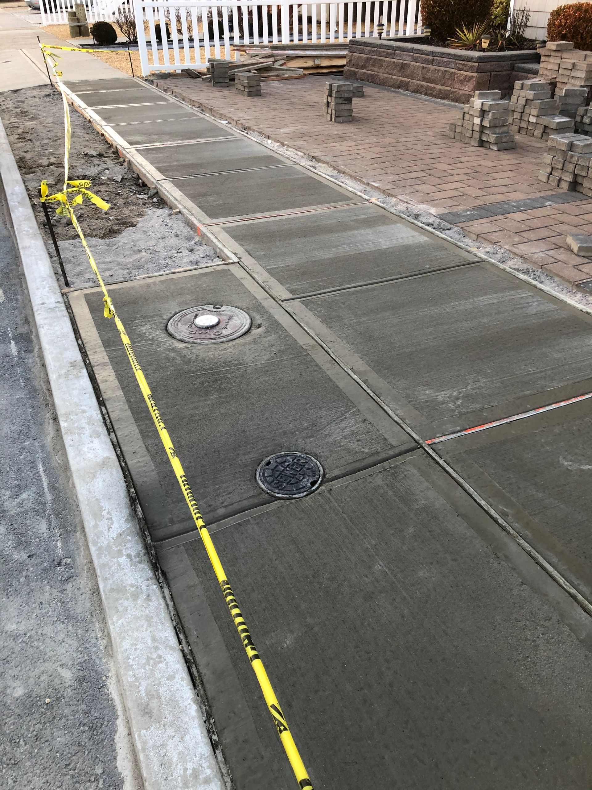 Newly poured concrete sidewalk with manhole covers, yellow caution tape along edges, construction site.