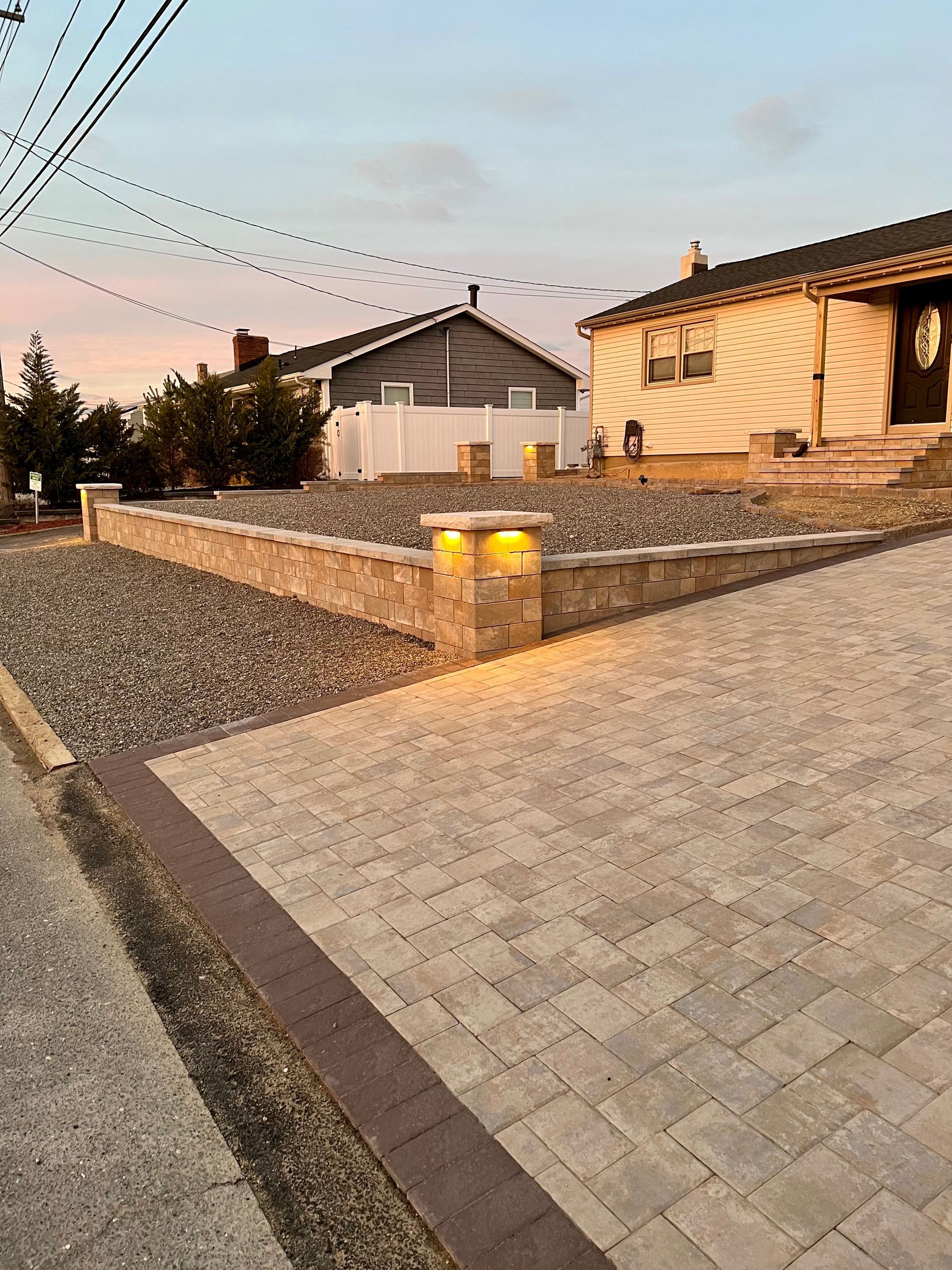 A paved driveway with a retaining wall, gravel bed, and illuminated pillars. Two houses in the background.