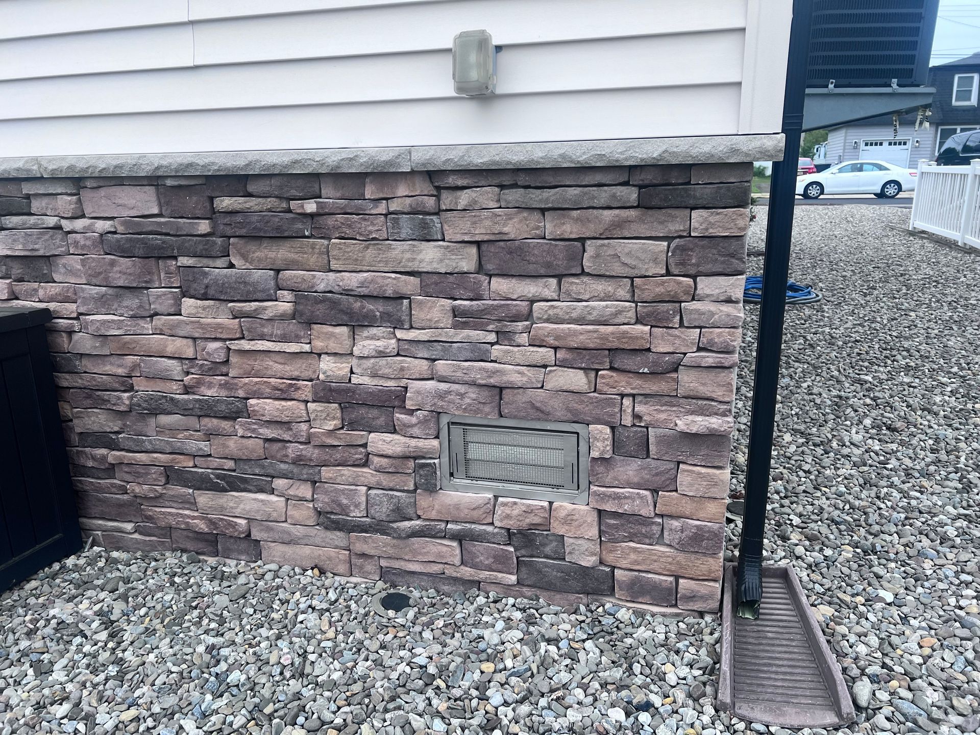 Stone-clad house foundation with vent, gravel ground, and black downspout.