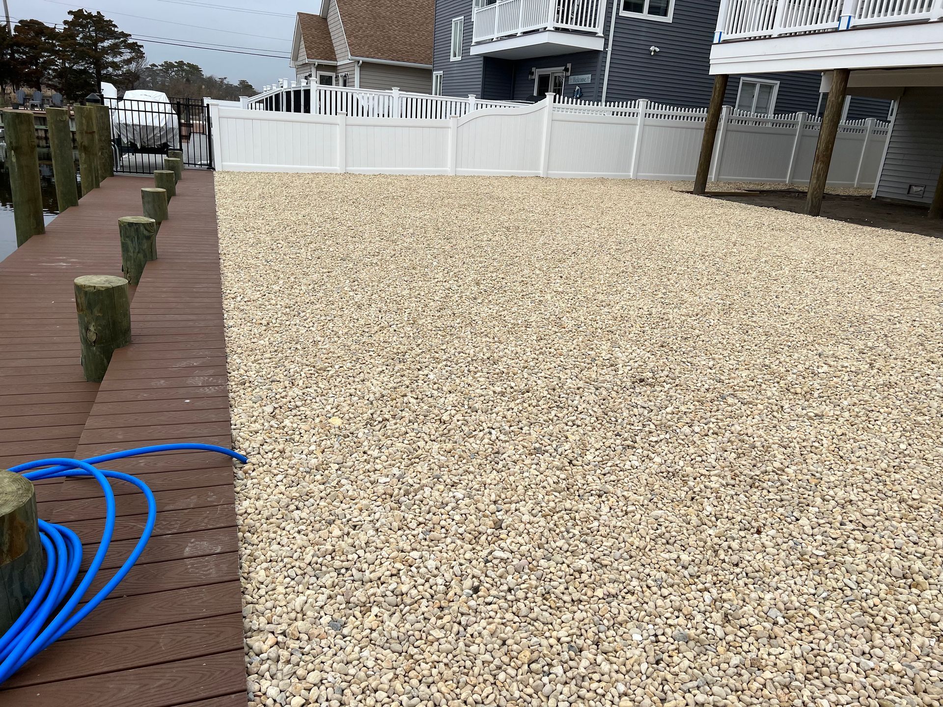 Dockside area with gravel ground, white fence, and surrounding houses. Blue hose on dock.