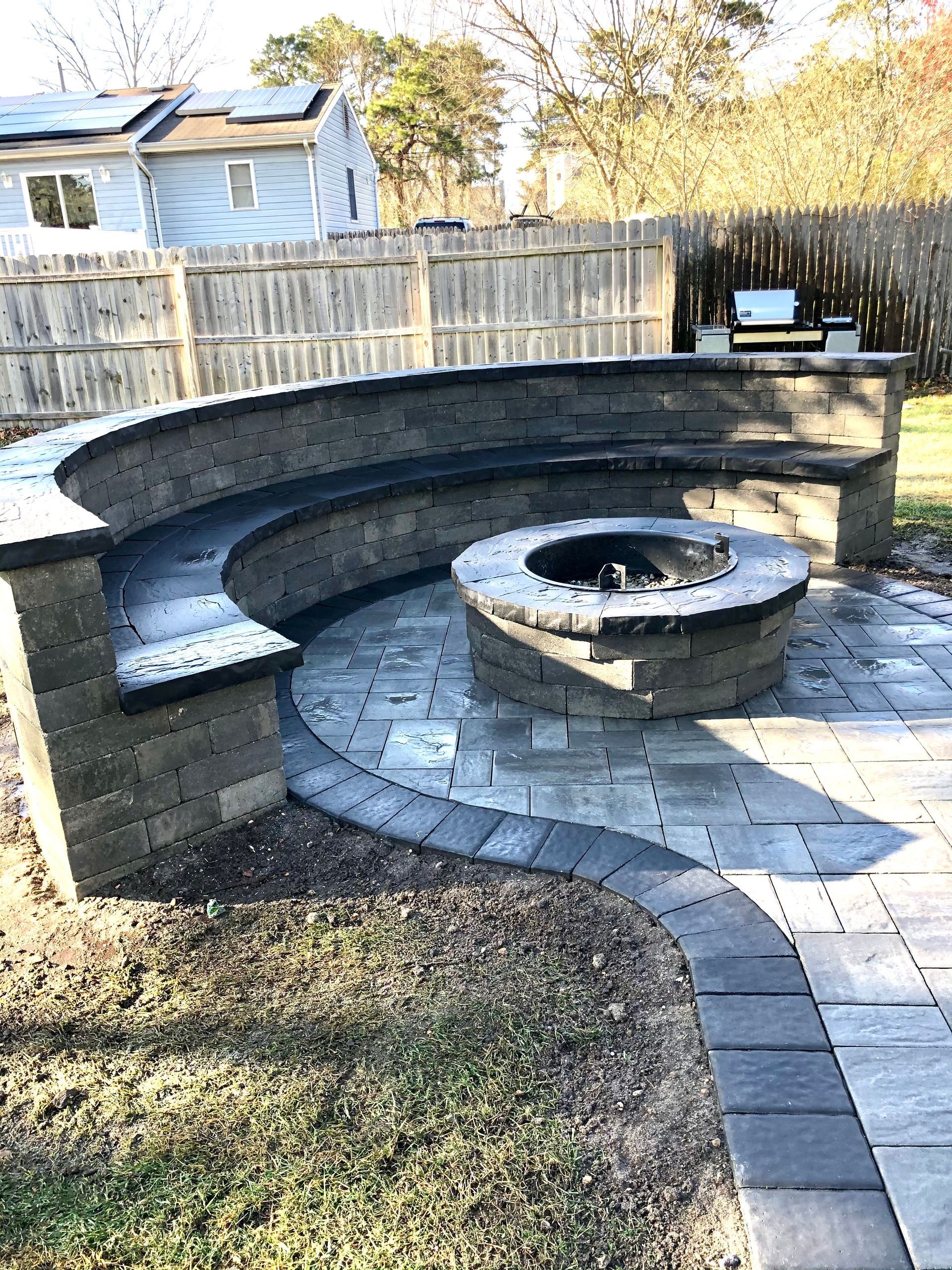 Stone fire pit with curved built-in seating on a paved patio, near a wooden fence and house.
