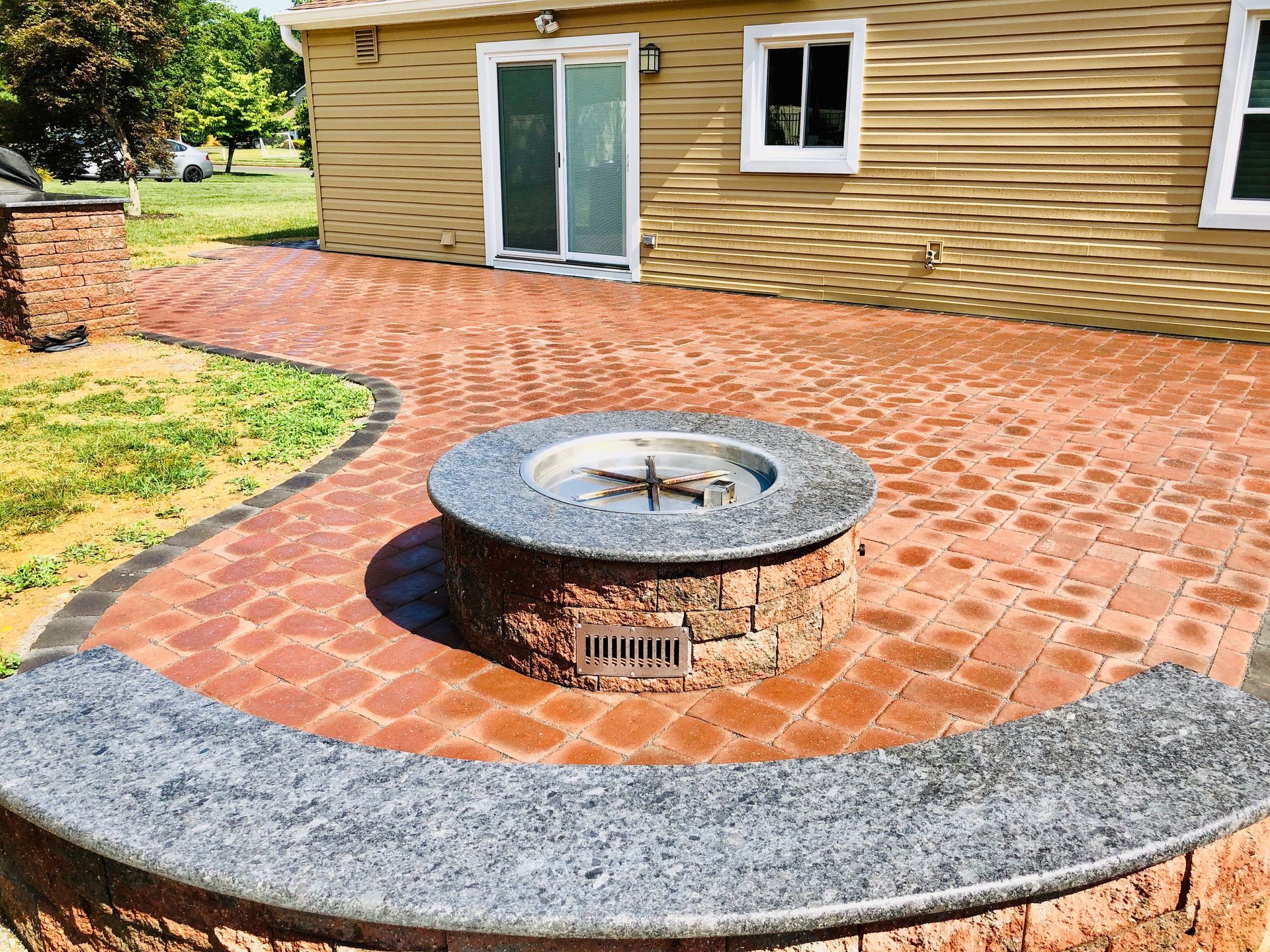 Brick patio with fire pit, bordering lawn. Tan house with sliding door and windows in the background.