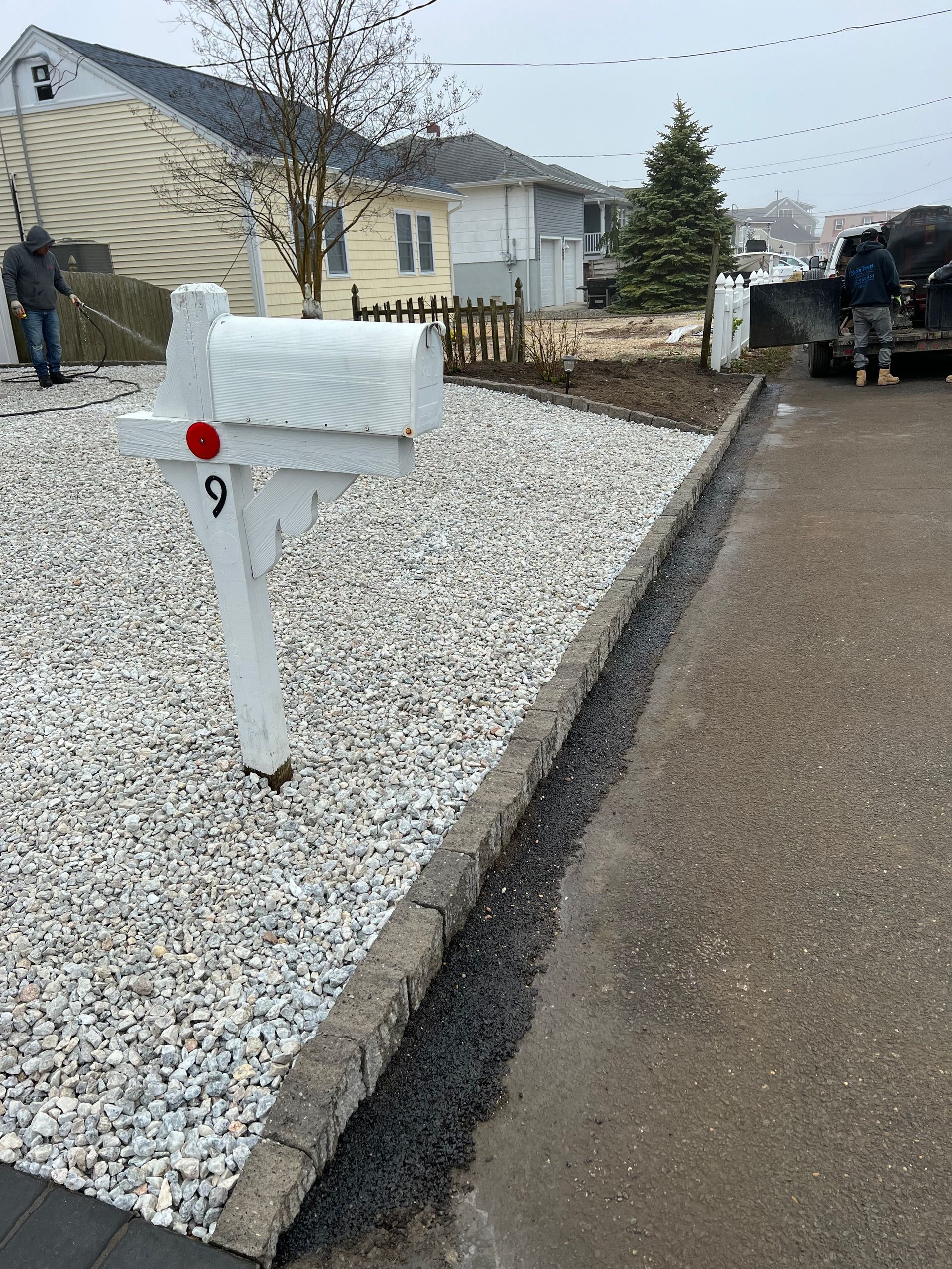 White mailbox on gravel bed next to driveway. Workers in the background. Cloudy day.
