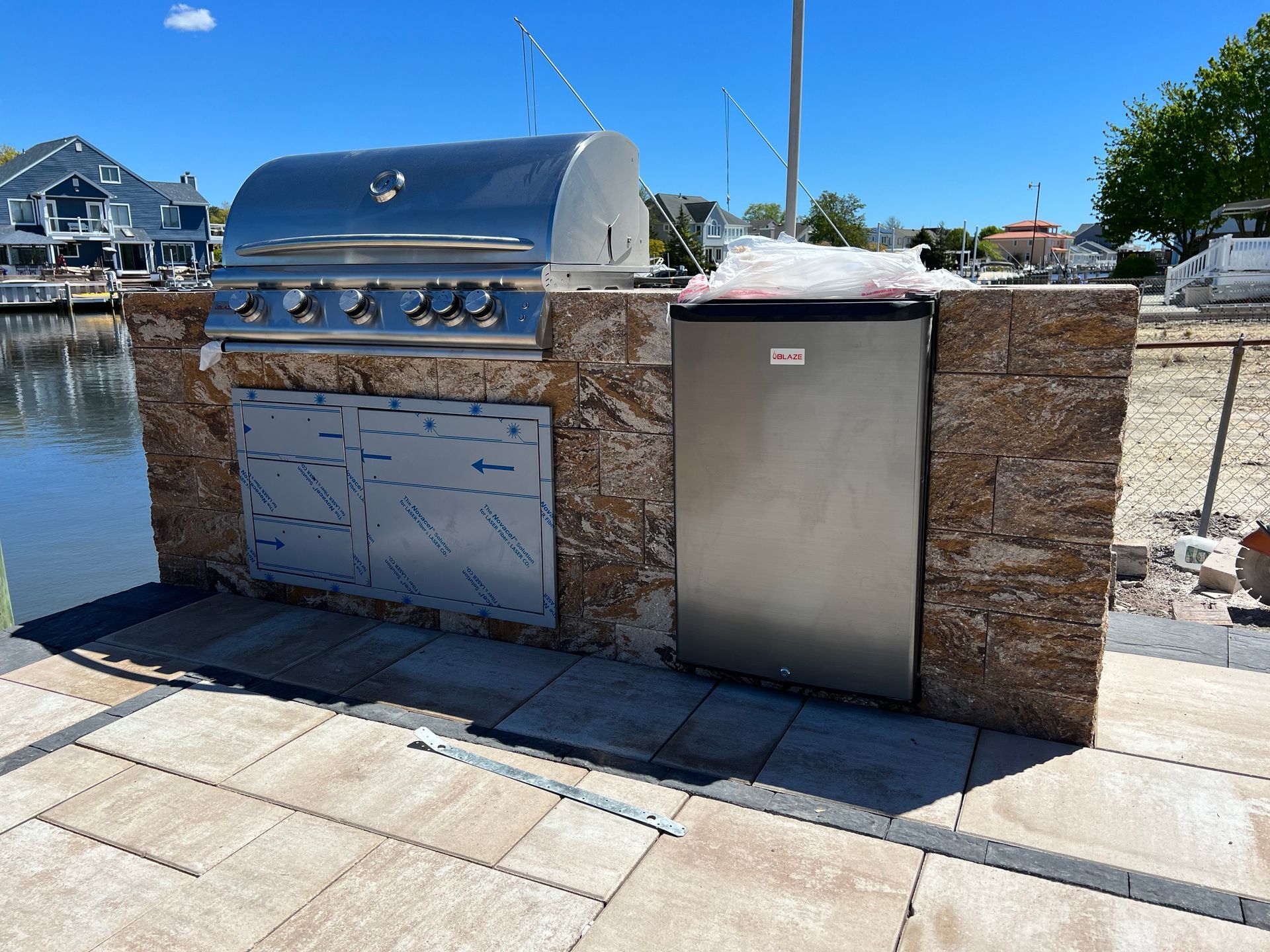 Outdoor kitchen with stainless steel grill, fridge, and stone surround, waterfront setting.