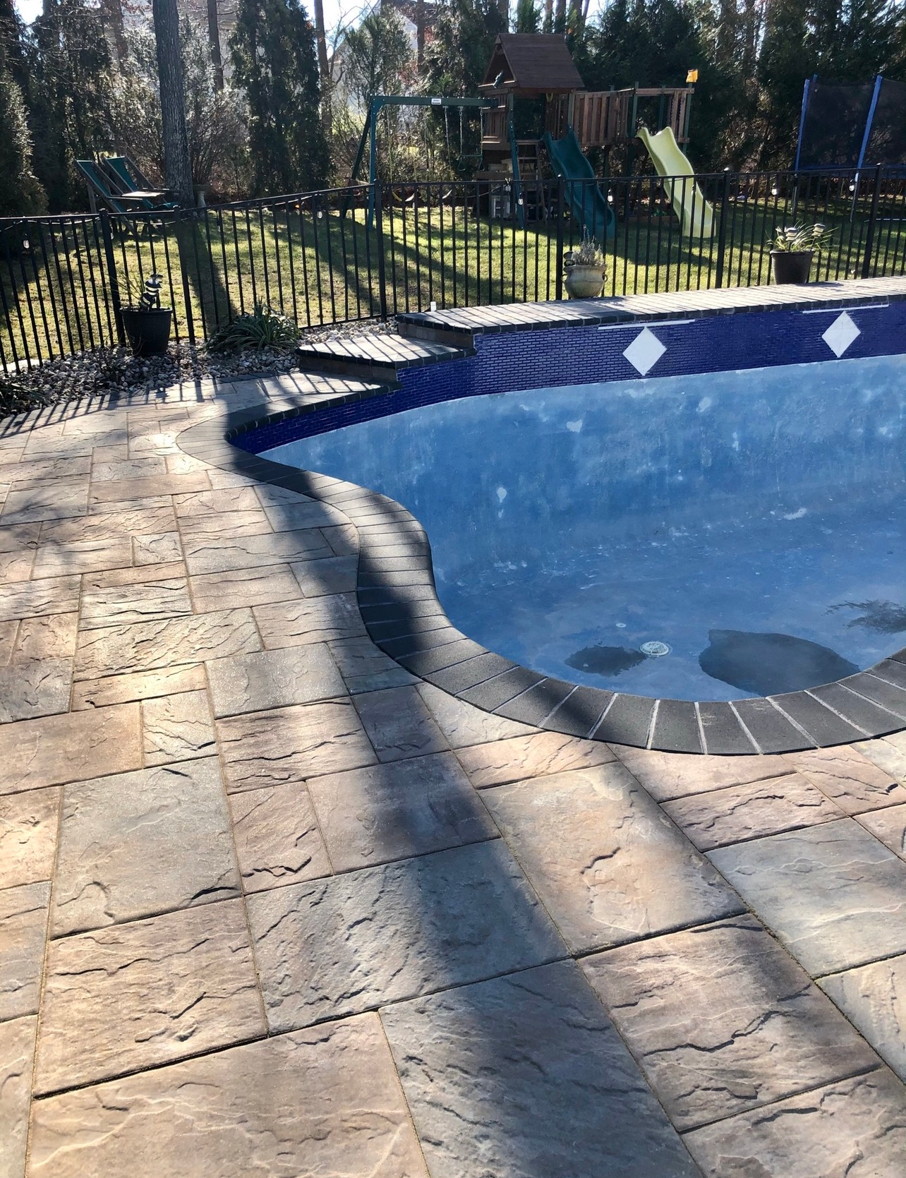 Poolside patio with a pool of blue water, surrounded by tan pavers and a black fence in a sunny outdoor setting.