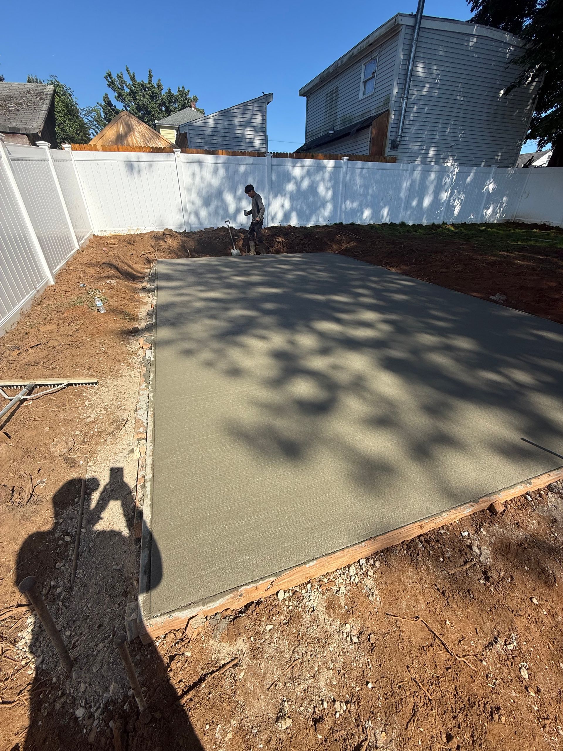 Concrete slab in backyard, worker smoothing surface. Dirt surrounds the slab, white fence in background.