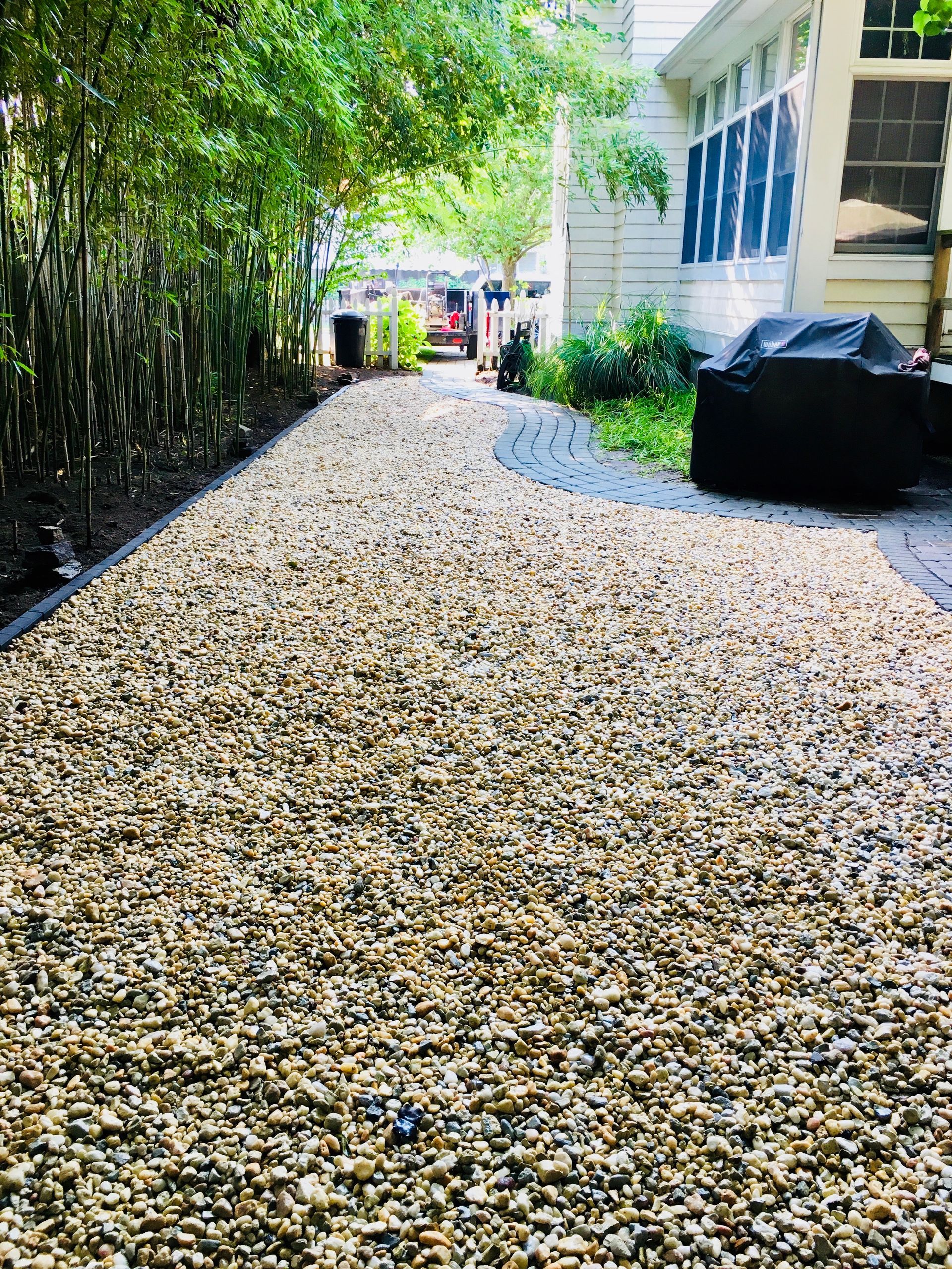 Gravel pathway leads past bamboo and a building to a patio area.