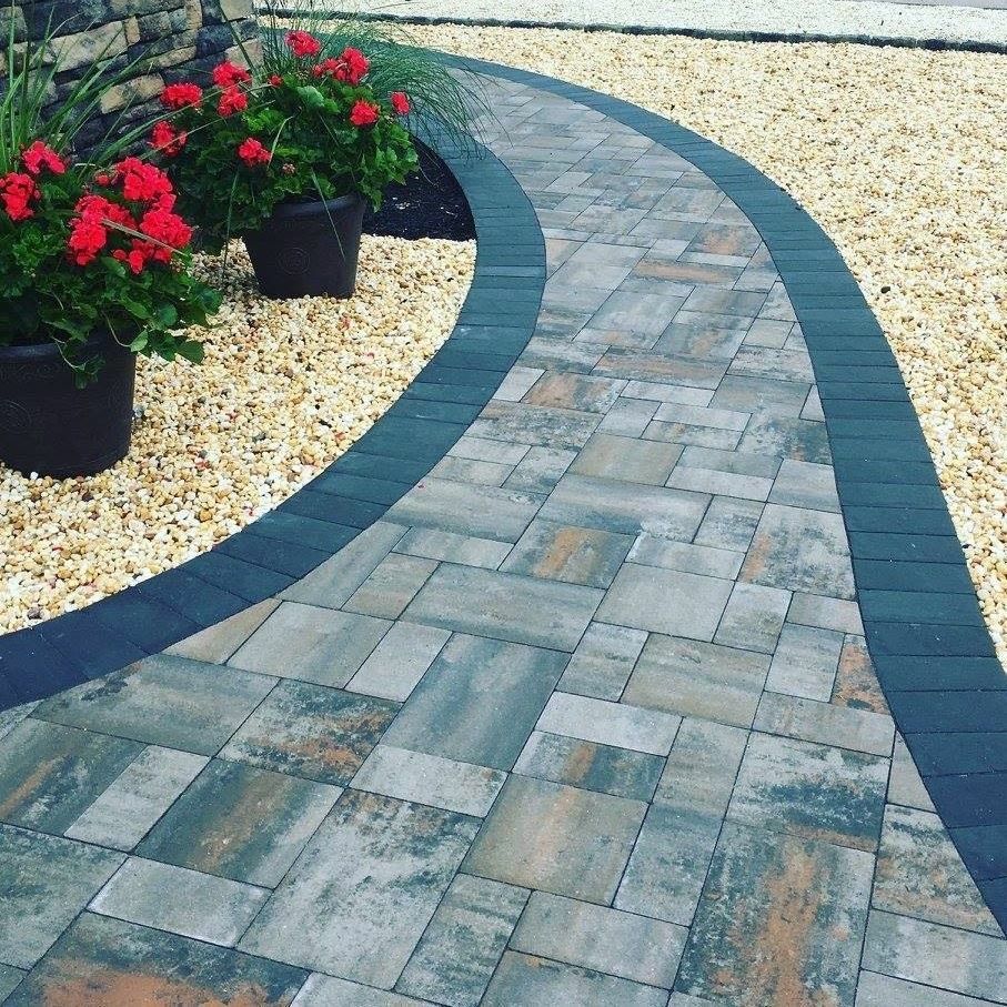 Curved paved walkway with dark border, gravel bed, and potted red flowers.