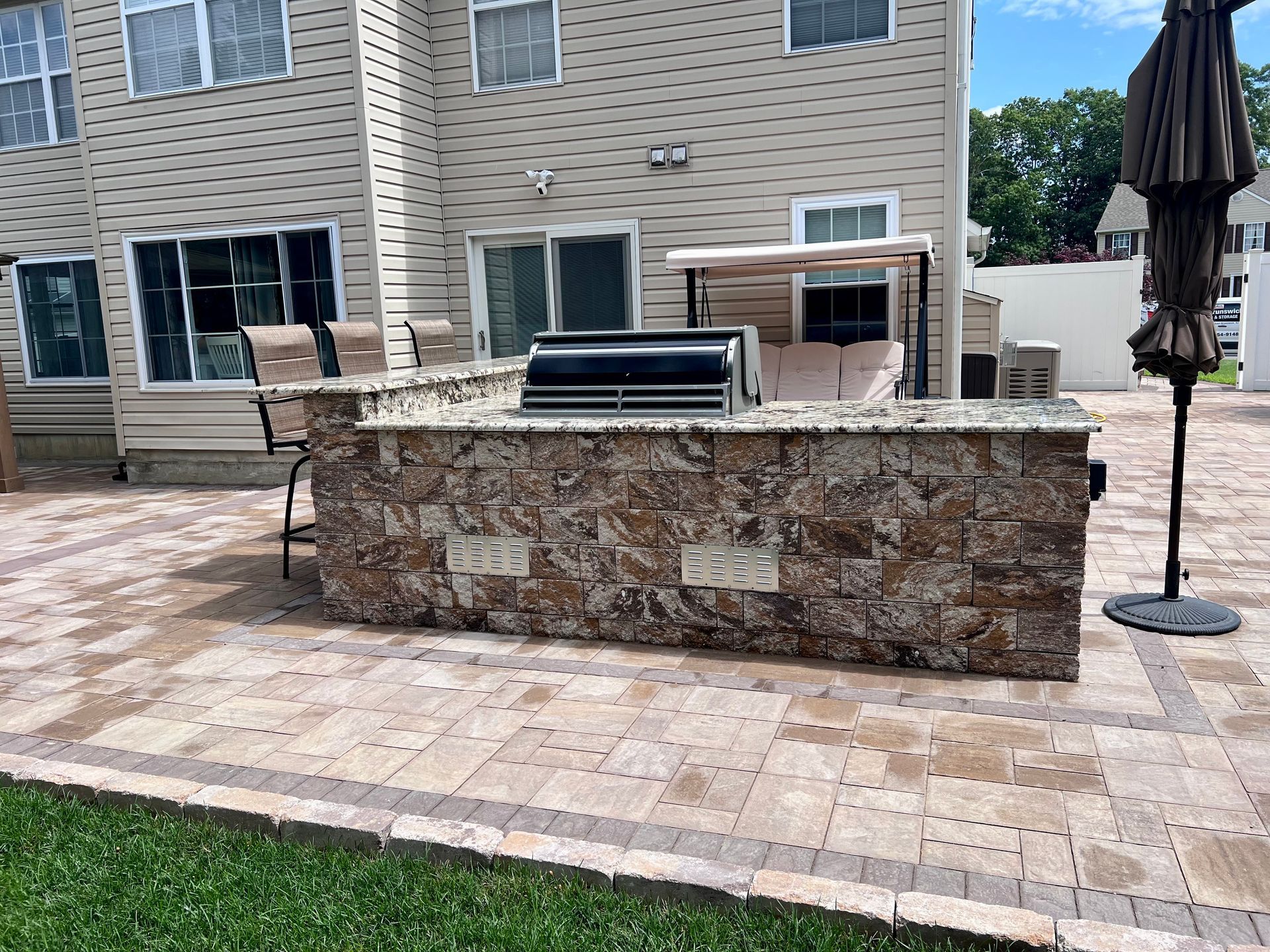 Outdoor kitchen with stone facade and grill on a brick patio in front of a house.