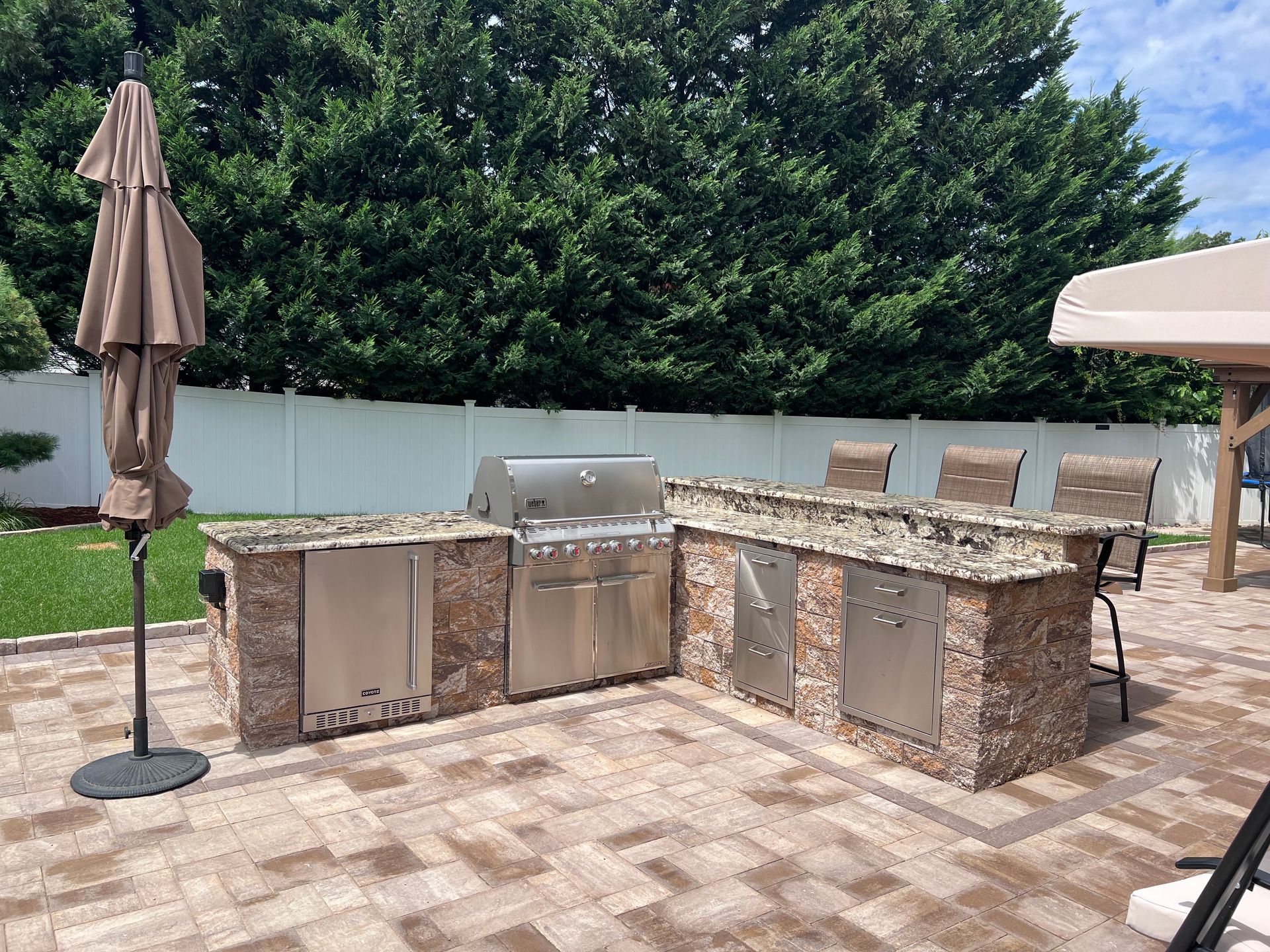 Outdoor kitchen with a grill, cabinets, and a bar with chairs on a brick patio.