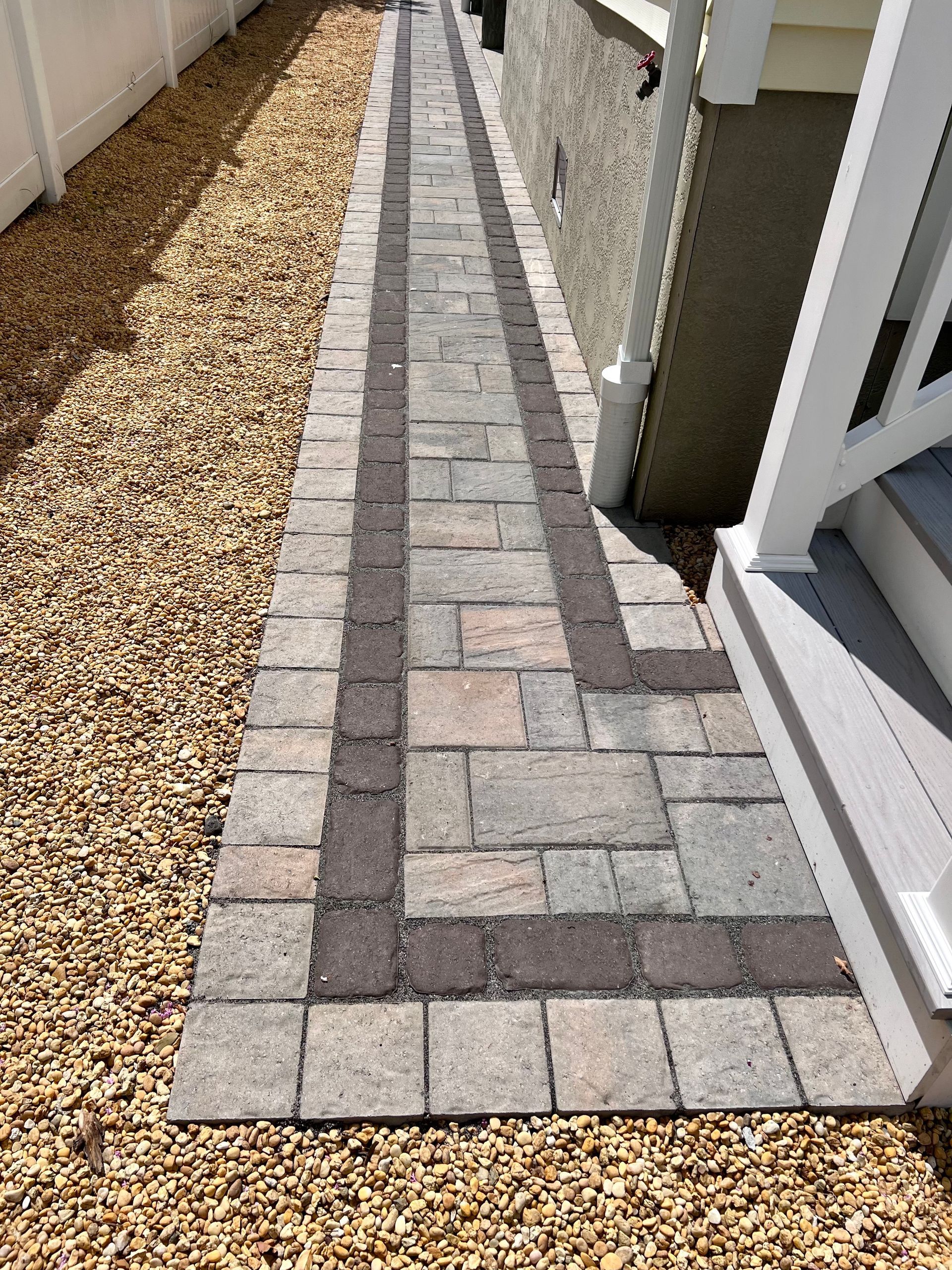 Brick pathway with brown and gray pavers, bordered by small brown pebbles, alongside a white fence and a house.