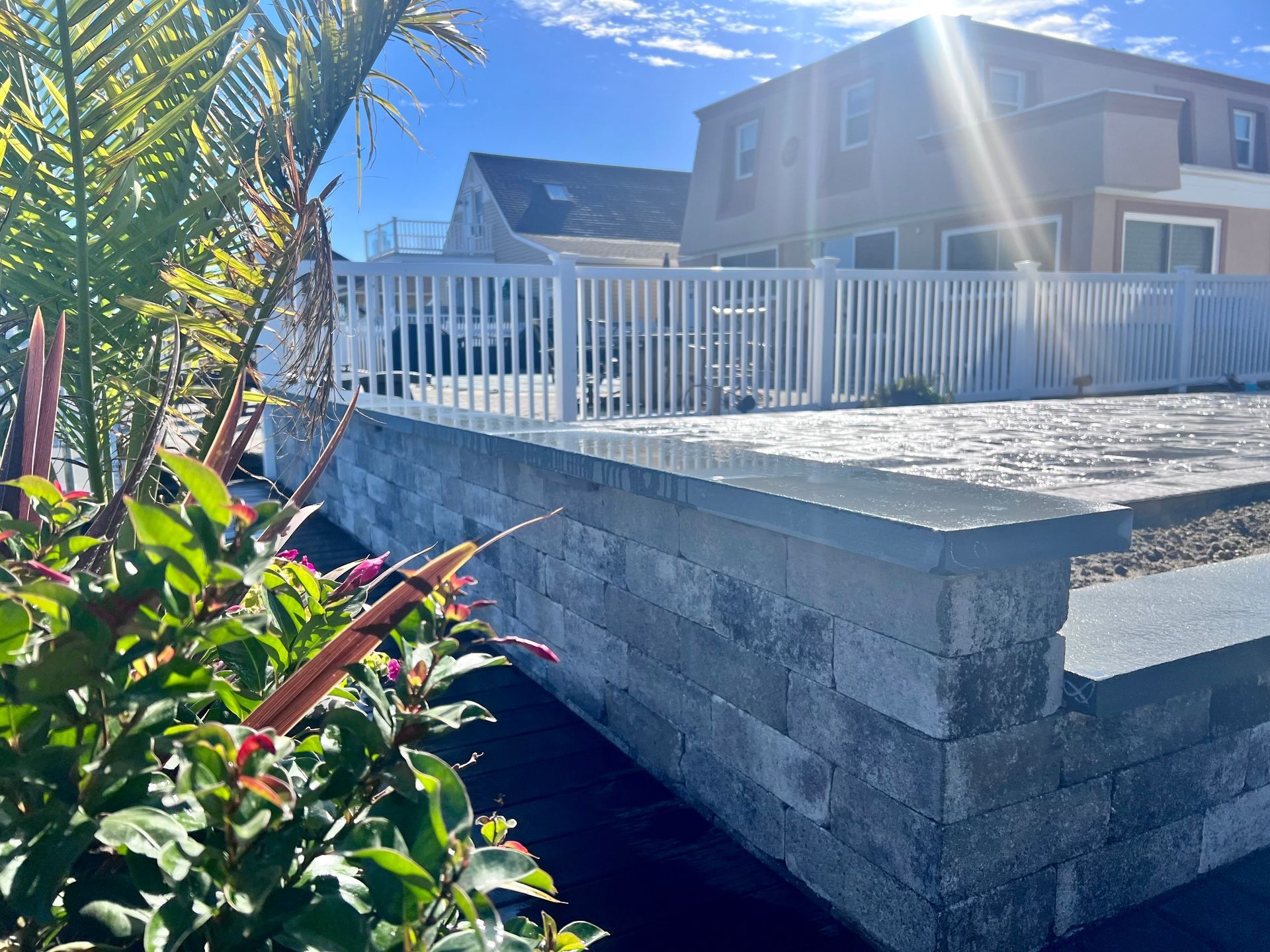 Gray block retaining wall with a white fence, home, and landscaping in sunny outdoor setting.