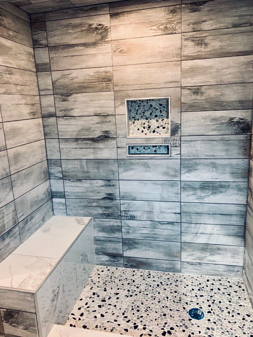 Shower interior with wood-look tile walls, a built-in bench, and a terrazzo floor. A small shelf in the wall holds decorative tiles.