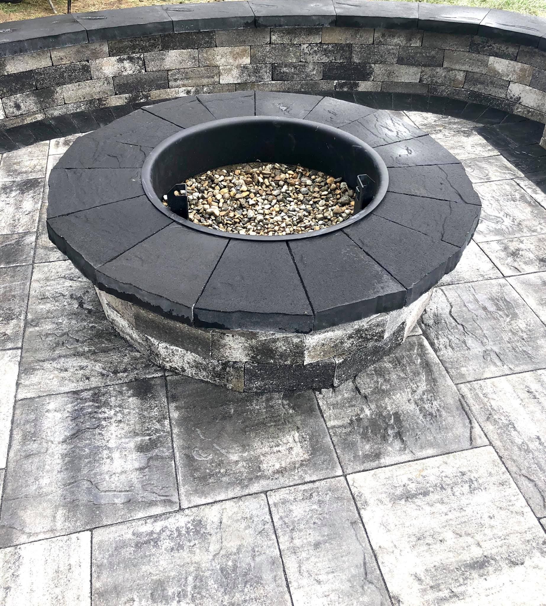 Circular fire pit with black rim and gray stone base, surrounded by a paved patio.