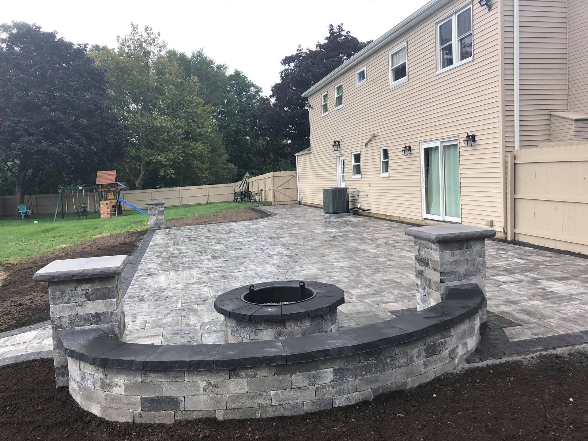 A newly constructed gray stone patio with a fire pit, next to a two-story beige house and backyard.