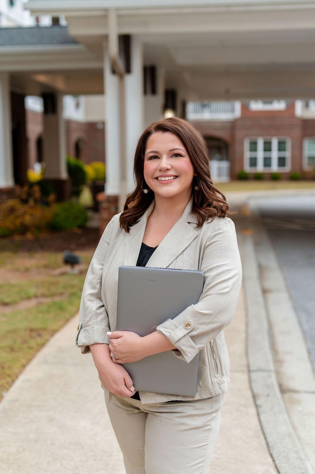 Woman in light suit smiles, holding a gray folder outside a brick building with white columns.