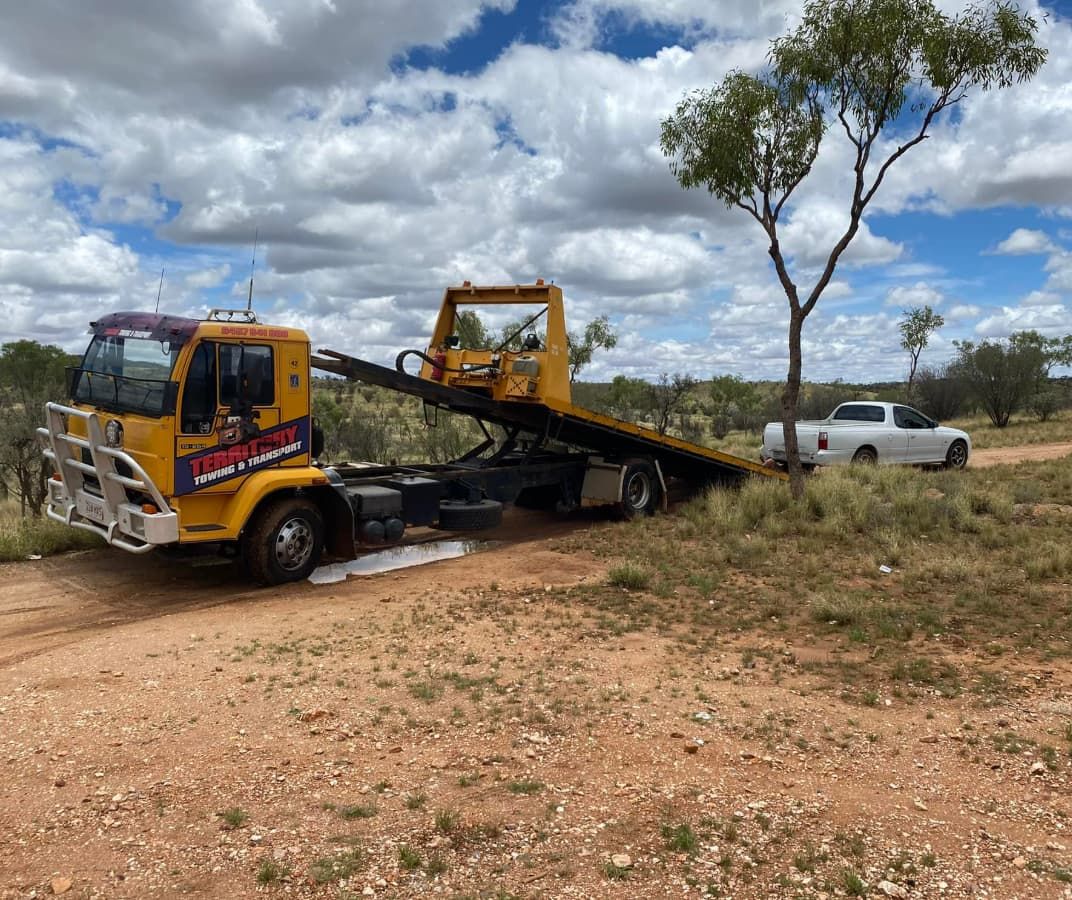 A Yellow Tow Truck Is Towing A White Truck On A Dirt Road — Territory Towing & Transport In Alice Springs, NT