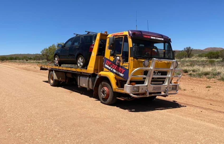 A Yellow Tow Truck Is Carrying A Black Car On A Dirt Road — Territory Towing & Transport In Alice Springs, NT