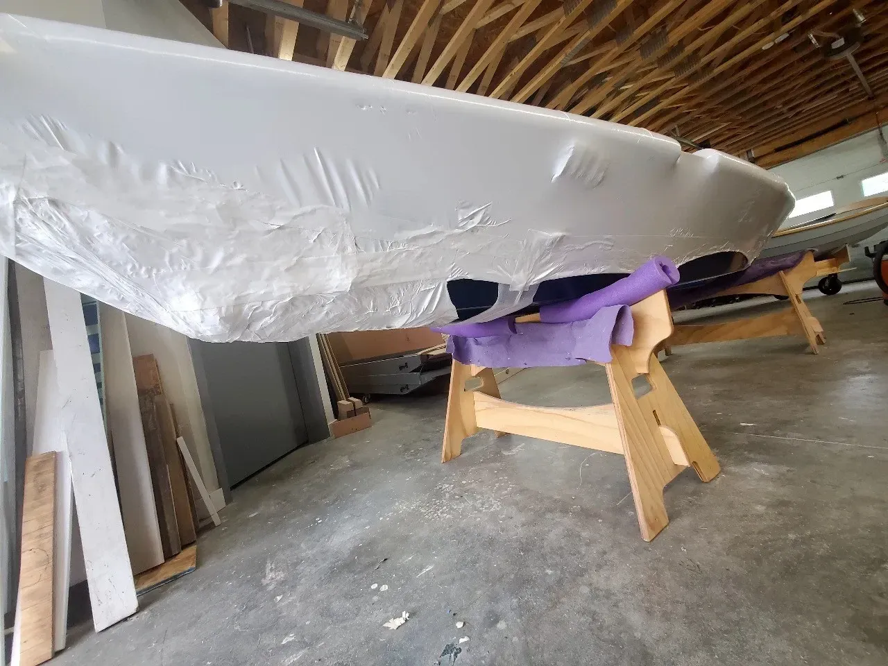 A boat is wrapped in plastic and sitting on a wooden stand in a garage.