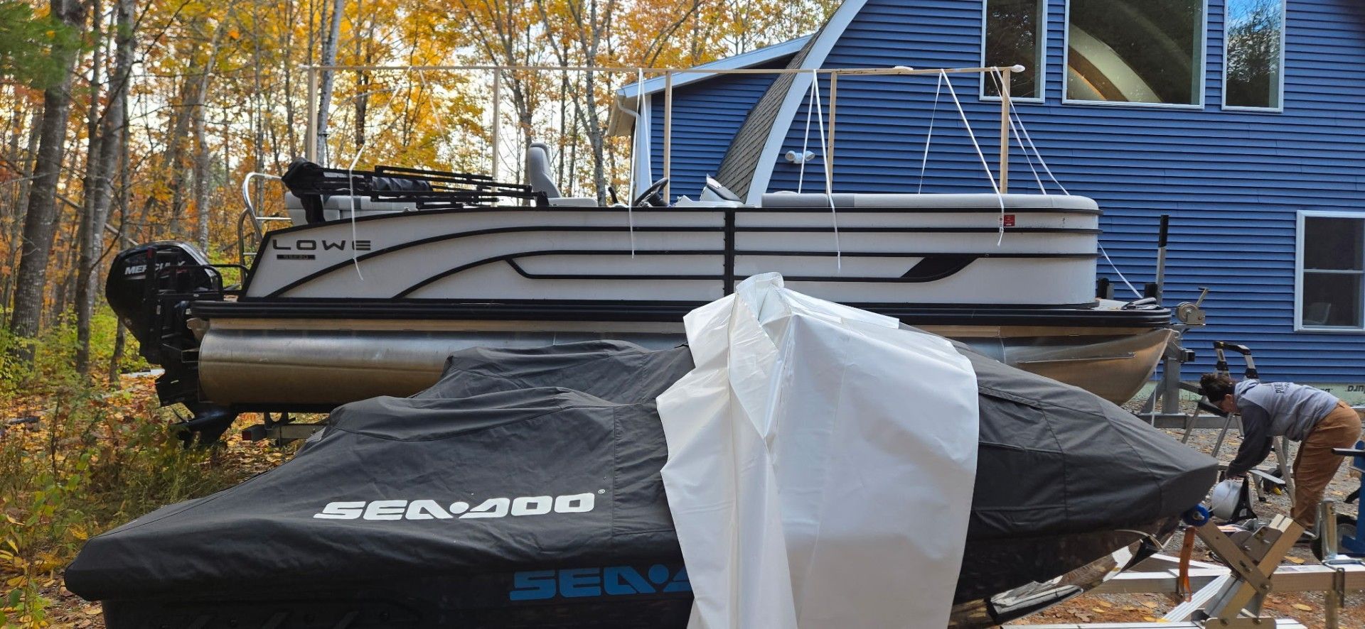 A pontoon boat and jet ski covered in tarps are next to a blue house, someone is working nearby.