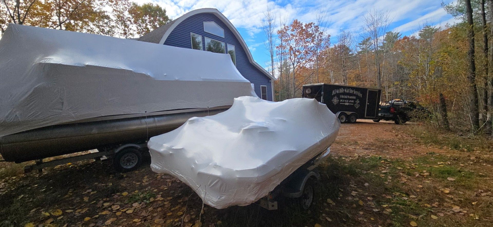 Boats covered in white shrink wrap sit in a yard near a blue barn and a black truck.