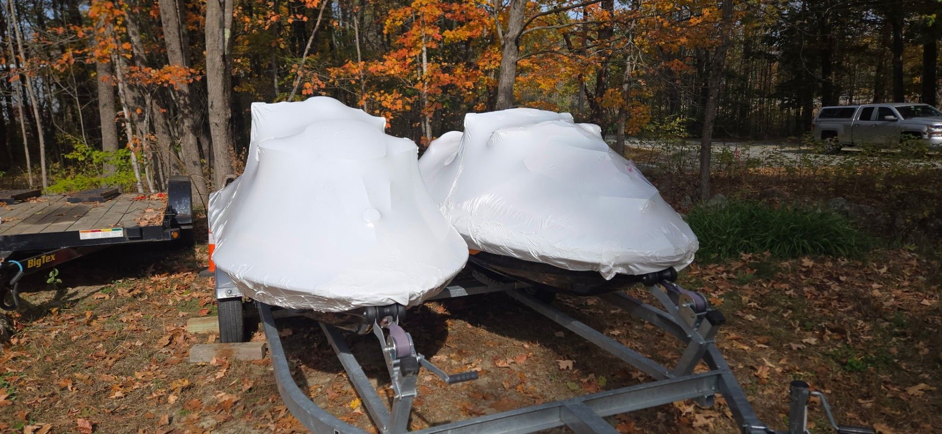 Two wrapped boats on a trailer in front of autumn trees.