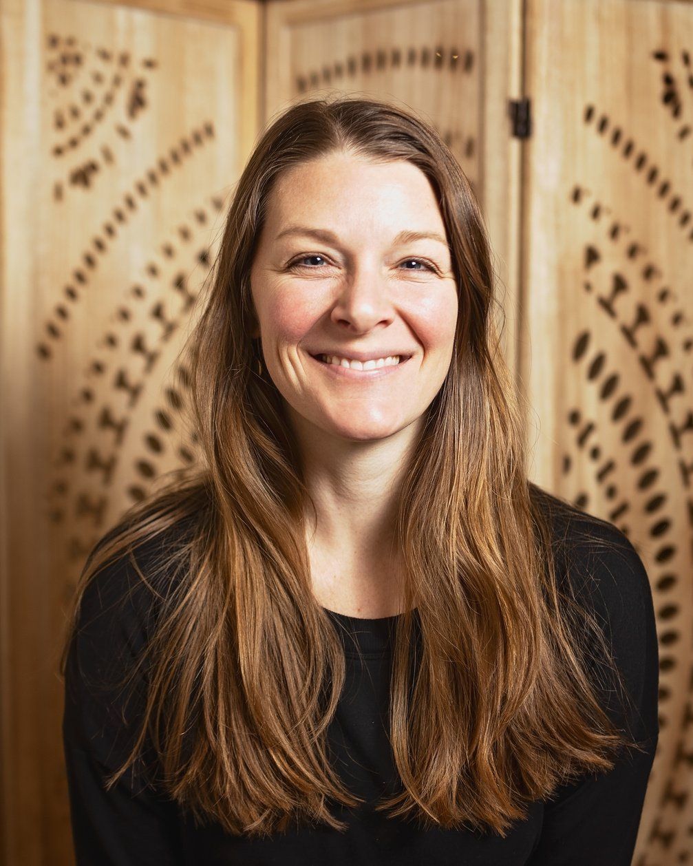 A woman with long brown hair is smiling in front of a wooden screen.