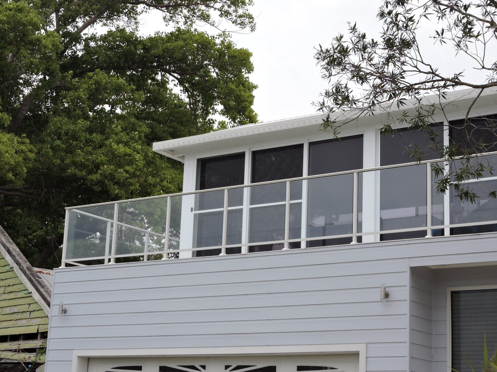 Gray house with glass and metal balcony railing; trees in background.