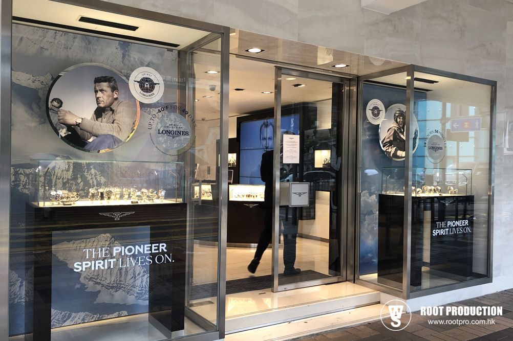 Exterior of a watch store. Glass windows display timepieces. A person enters through the open door.