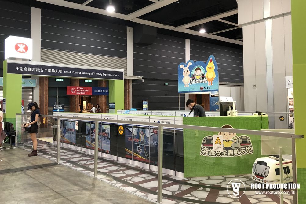 An indoor train station scene, with green and white details, a person in a booth, and a woman waiting.