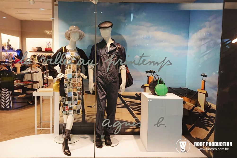 Store window display with mannequins wearing clothes against a painted background and a white pedestal.