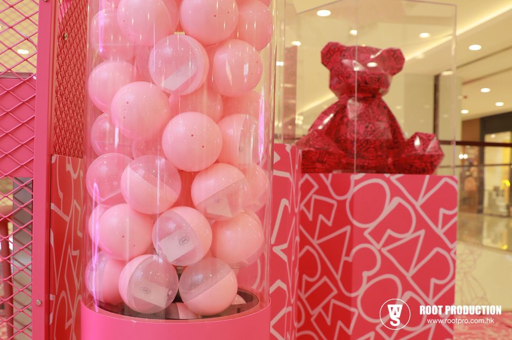 Pink gumball machine with prizes, next to a red bear statue on a patterned pink pedestal, in a mall setting.